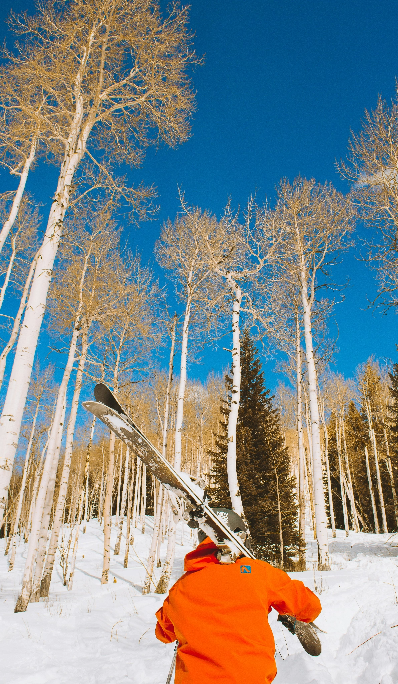 Person wearing orange snow suit carrying skis up snowy hill with trees