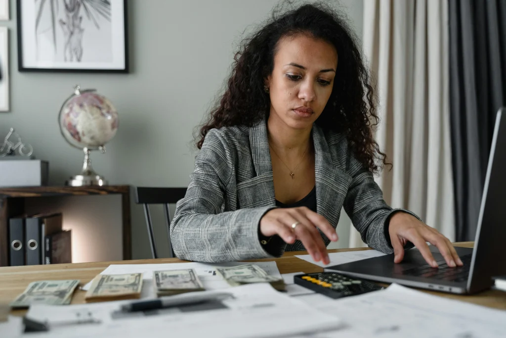 Woman wearing business suit jacket sitting at table with money and papers scattered, a calculator, and laptop open