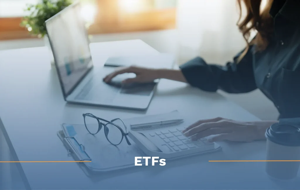 What is an ETF? Woman sitting at desk with laptop open, calculator, glasses, and clipboard