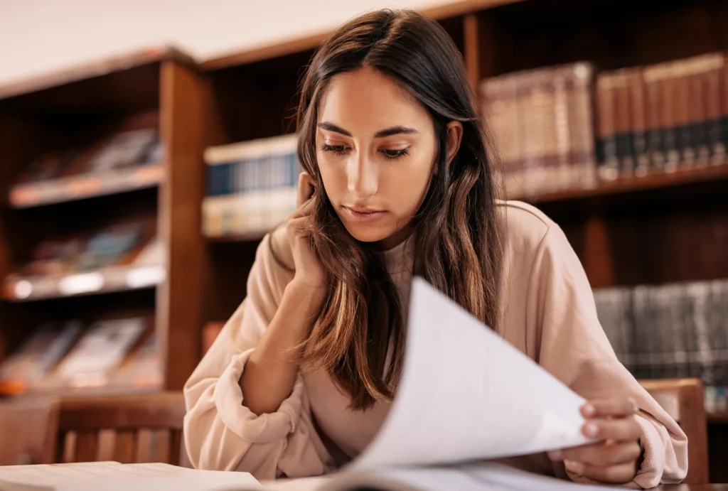 Young woman sitting in desk at library flipping through papera
